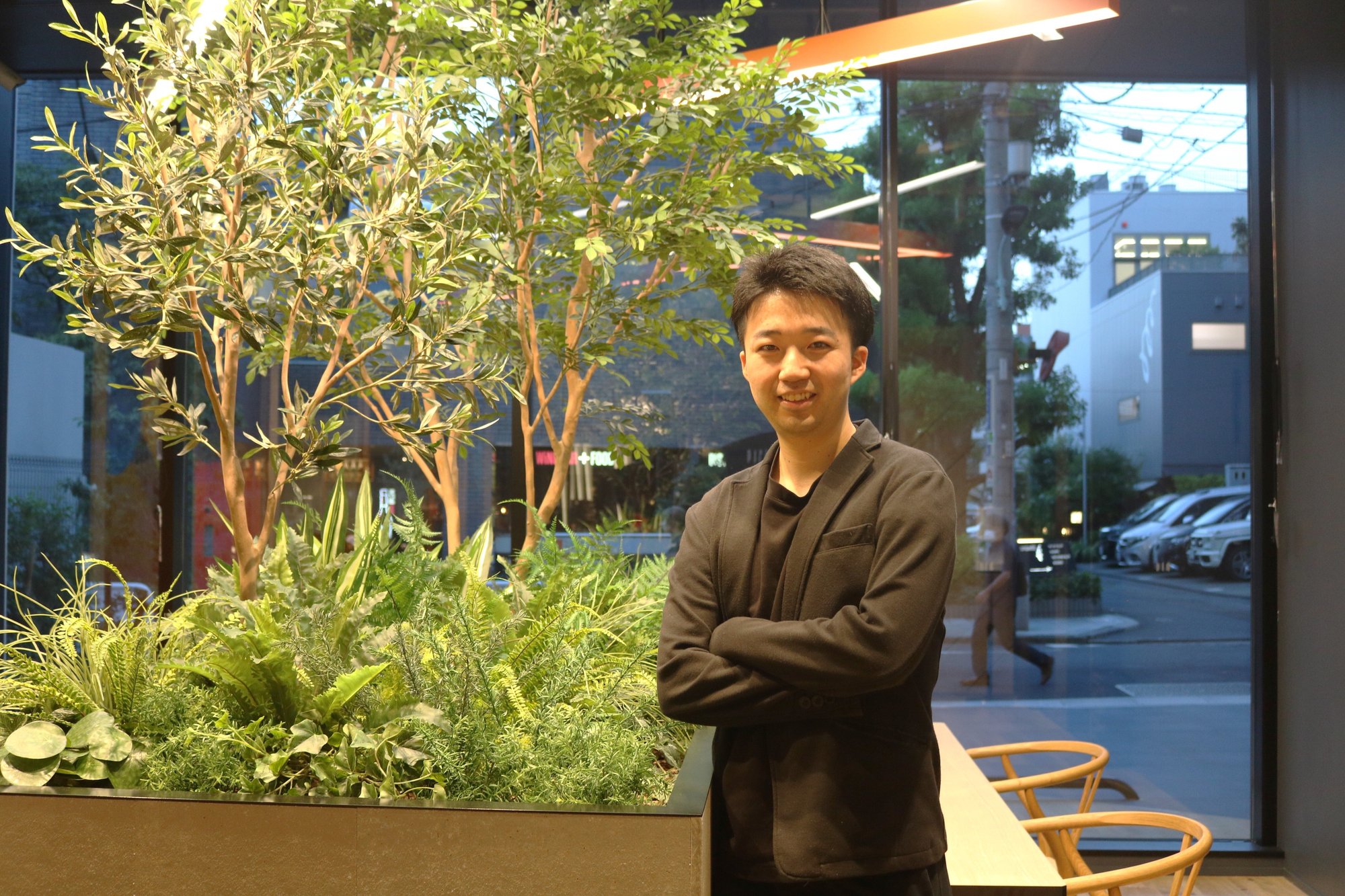 屋外の植栽の前で腕を組んで立つNさん