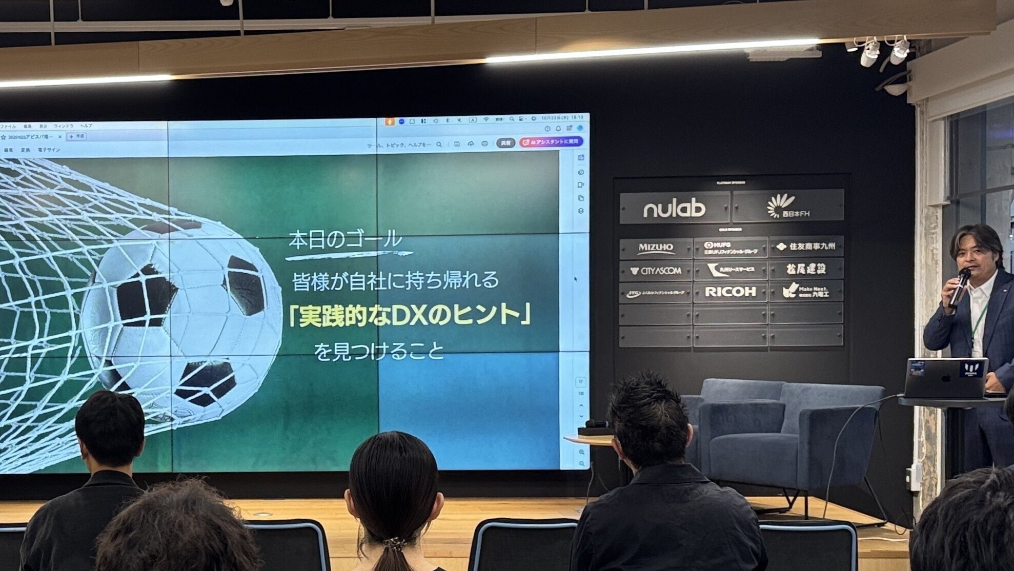 スクリーンに「本日のゴール 皆様が自社に持ち帰れる『実践的なDXのヒント』を見つけること」と映し出された会場の様子。登壇者がマイクを持って話している