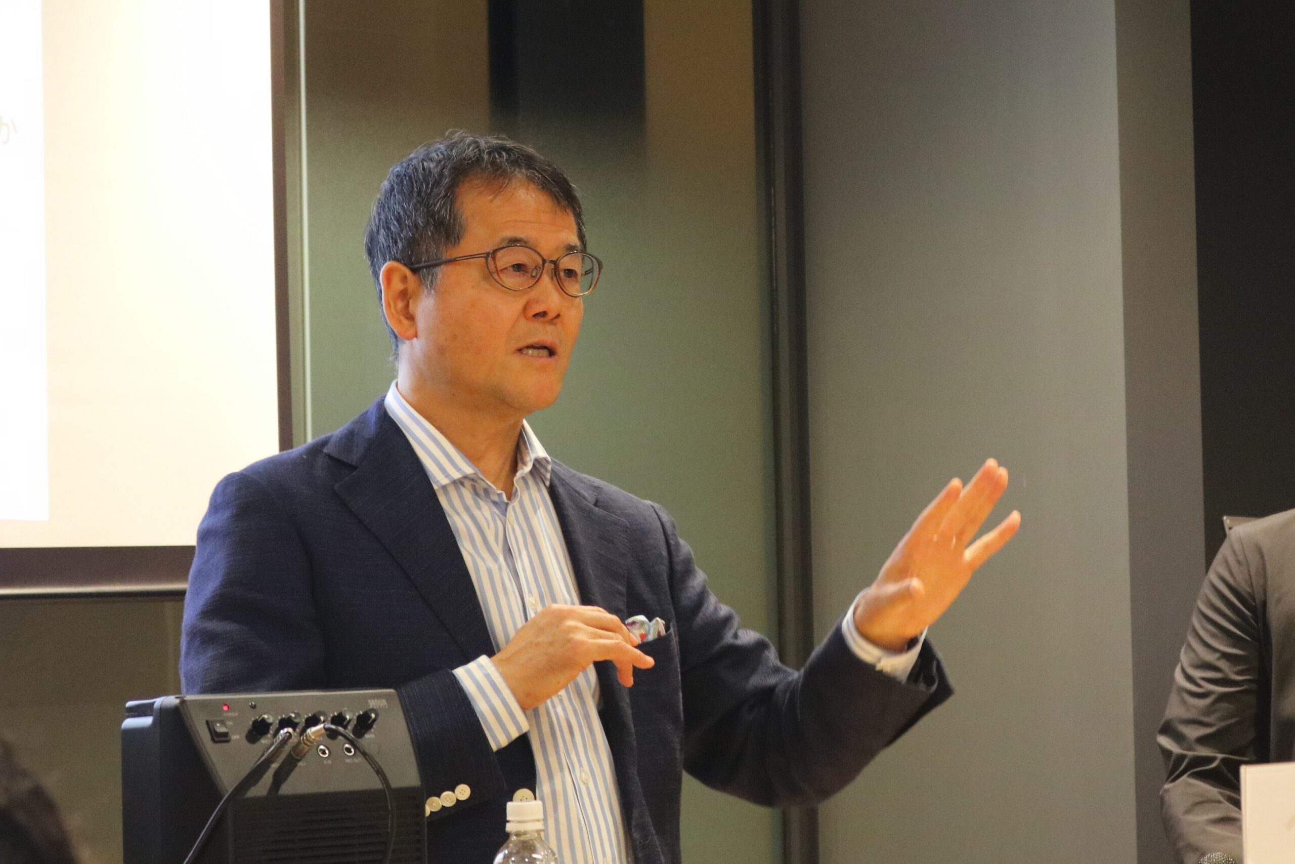 スクリーンを背に、手ぶりを交えて講演する菅野誠二氏