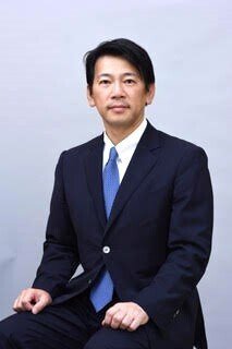 『瞬考』著者の山川隆義氏。紺のスーツに水色のネクタイで椅子に座ったポートレート